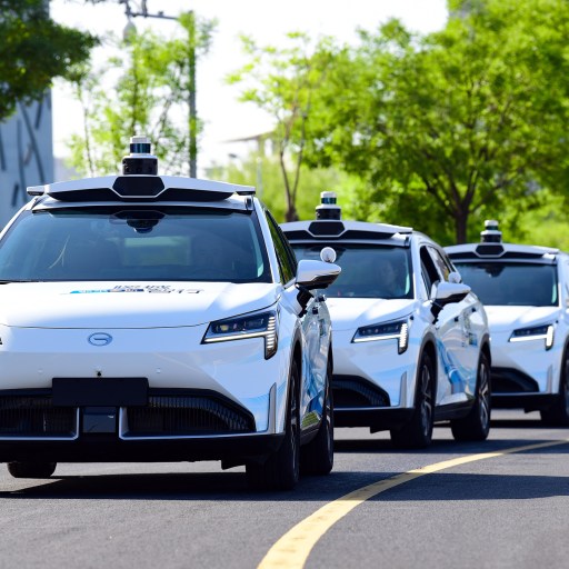 Three white autonomous cars drive down a city street lined with trees.