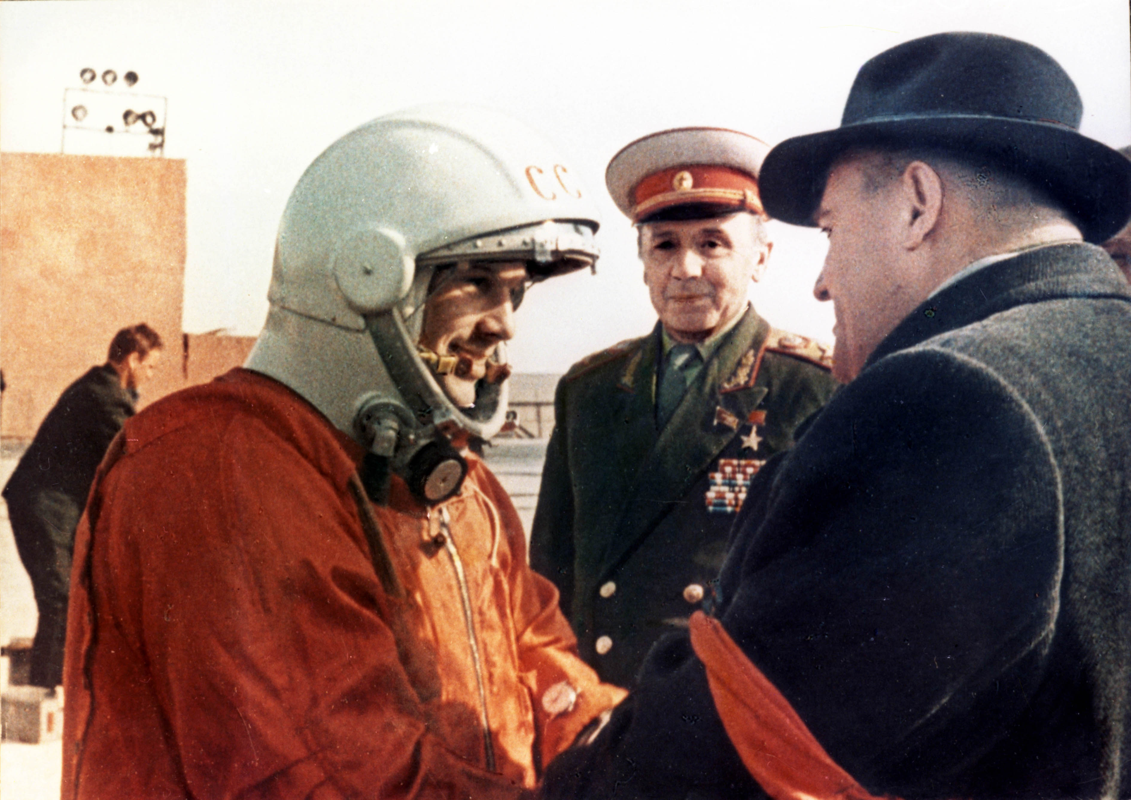 A man in a space suit, symbolizing the USA's lunar achievements, shakes hands with a man in a dark coat and hat, reminiscent of USSR diplomacy, while another man in a military uniform watches.