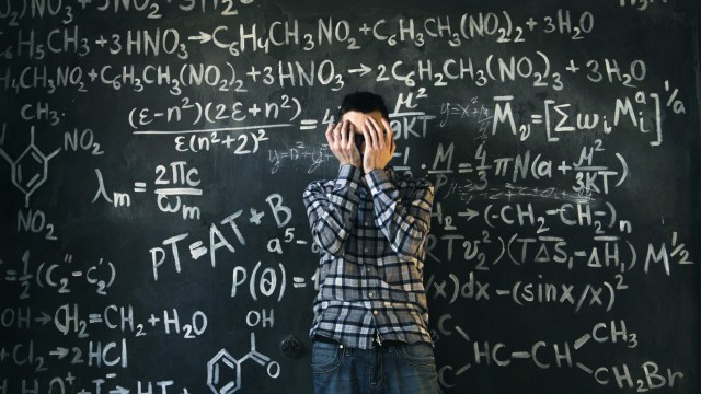 A person stands in front of a chalkboard filled with complex mathematical formulas, covering their face with their hands.