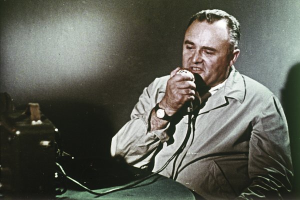 A man in a coat speaks into a microphone at a table with equipment, passionately discussing the historic moon landing that marked a pivotal moment between the USA and USSR.