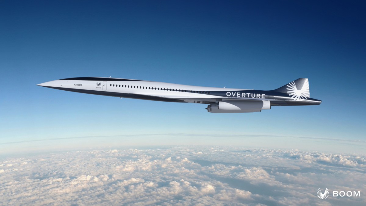 A sleek supersonic jet labeled "Overture" by Boom flies above the clouds against a clear blue sky.