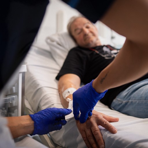 A healthcare worker wearing blue gloves gently inserts an IV into a man's hand as he lies on a hospital bed, battling lung cancer.