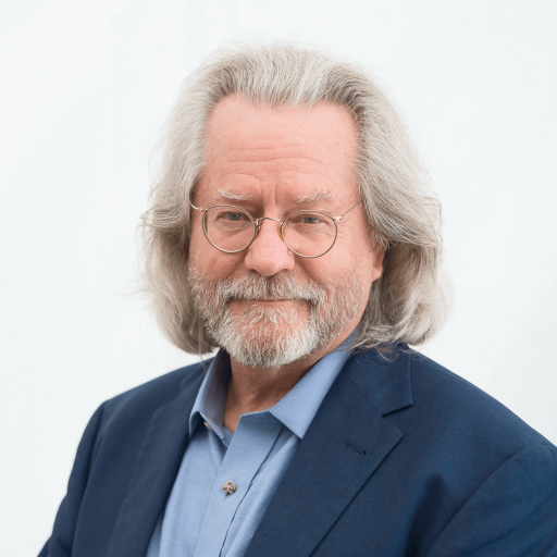 A person with long gray hair, glasses, and a beard, wearing a blue shirt and dark jacket, smiles slightly against a plain white background, exuding a serene aura reminiscent of religious tranquility.