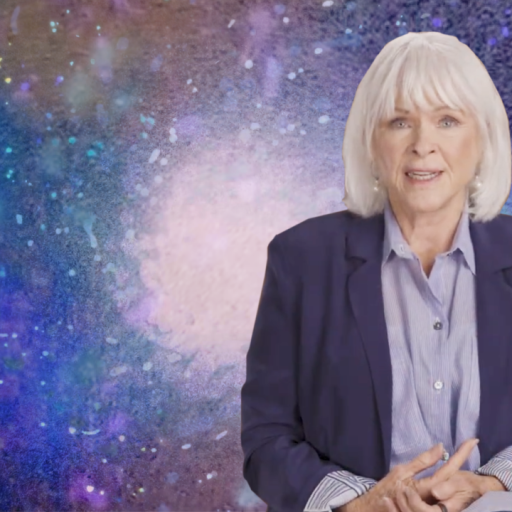A person with white hair sits in front of a colorful, abstract background. They are wearing a dark blazer over a striped shirt.
