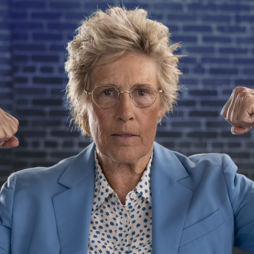 Person wearing a light blue suit and floral shirt, flexing both arms, set against a brick wall background.