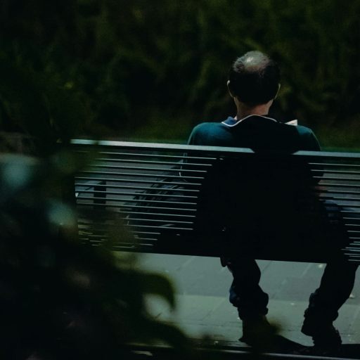 A lifelong single with short hair sits alone on a park bench at night, facing away. Bushes and dim lighting surround the scene.