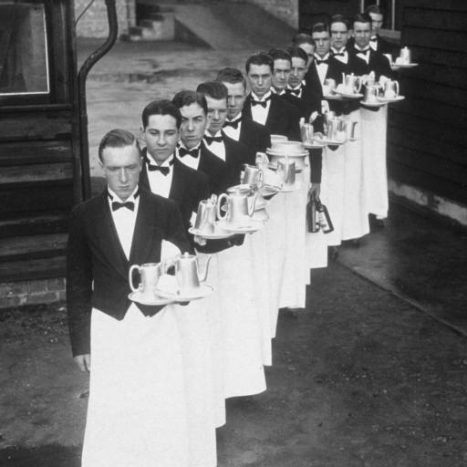 A line of waiters in formal attire, embodying a sense of unreasonable hospitality, carry trays with teapots and cups as they gracefully walk in procession outside the building.