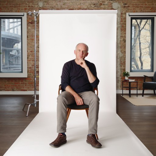 A person sitting on a chair in a cozy interior with brick walls, two windows, plants, and a lamp.