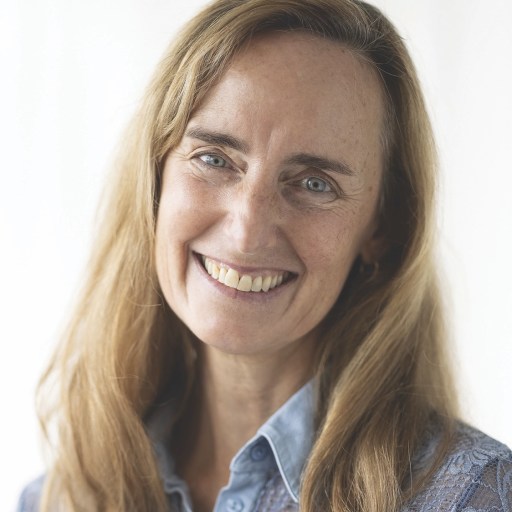 A woman with long hair smiles while wearing a blue blouse against a neutral background.