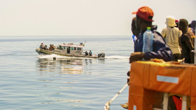 A group of people on a boat watch as a patrol vessel approaches on calm, open water under a clear sky, reflecting ongoing migration statistics in the region.