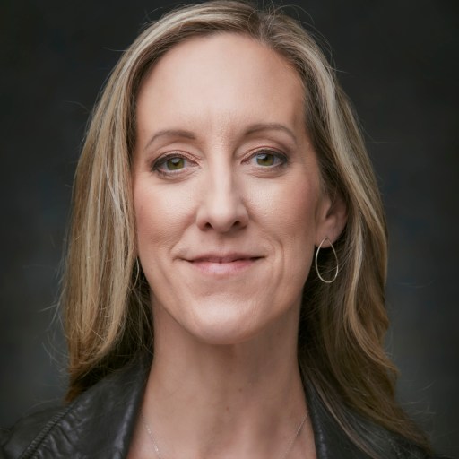 A person with long blonde hair and a leather jacket smiles slightly against a dark background.
