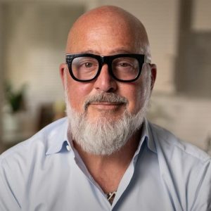 A person with a bald head, white beard, and black glasses is wearing a light blue shirt, standing in a softly lit kitchen.