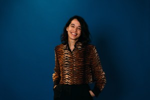 A person with dark hair smiles, wearing a tiger-striped shirt against a dark blue background.