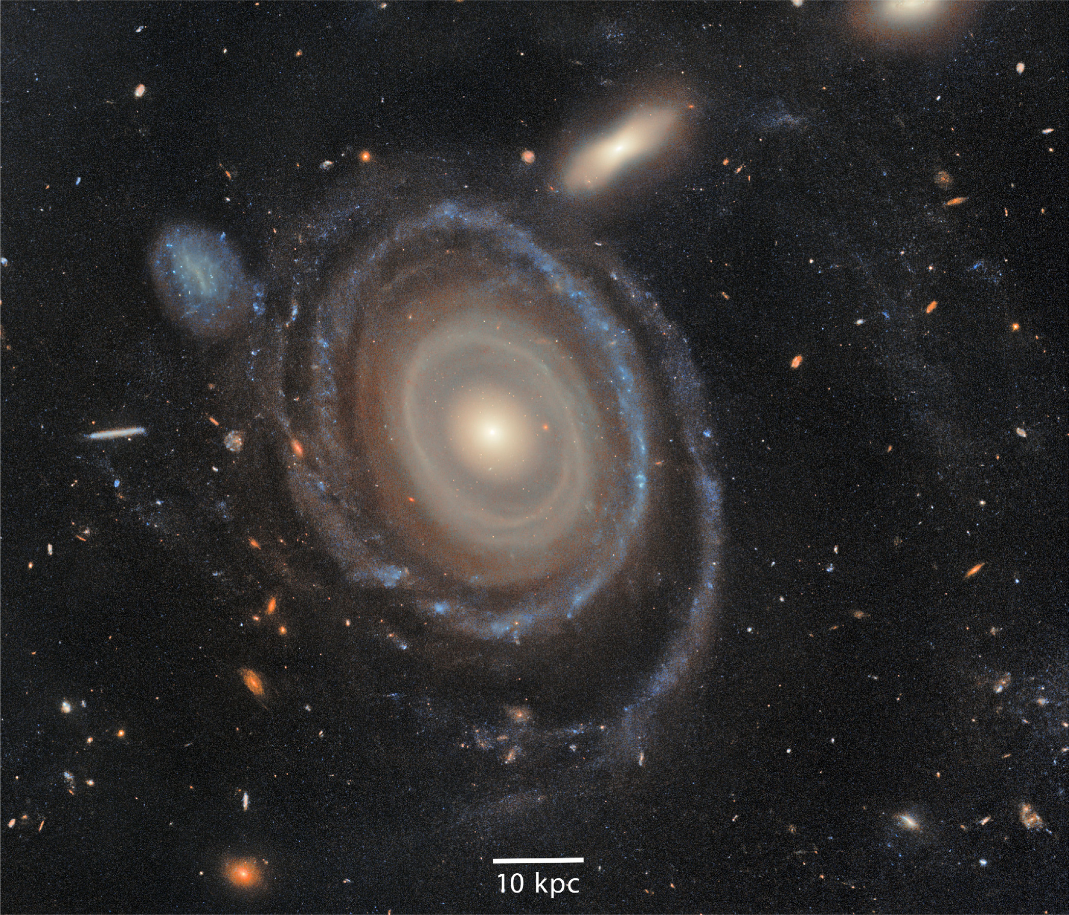 A spiral galaxy with a bright core and distinct arms, resembling a bullseye ring galaxy, is surrounded by smaller galaxies, stars, and cosmic dust. A scale labeled "10 kpc" graces the bottom center.