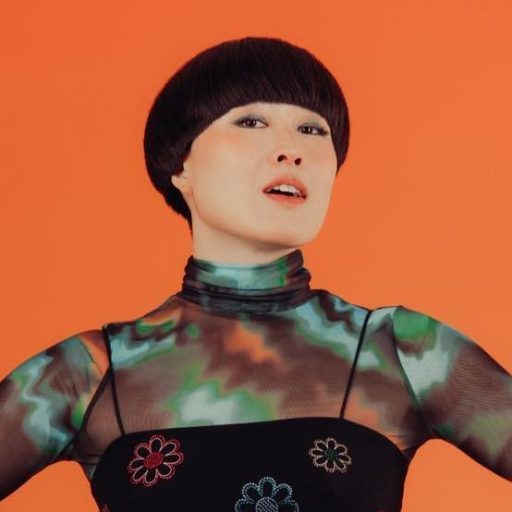 Person posing against an orange background, wearing a green and black patterned top with flower designs and high-waisted green pants.