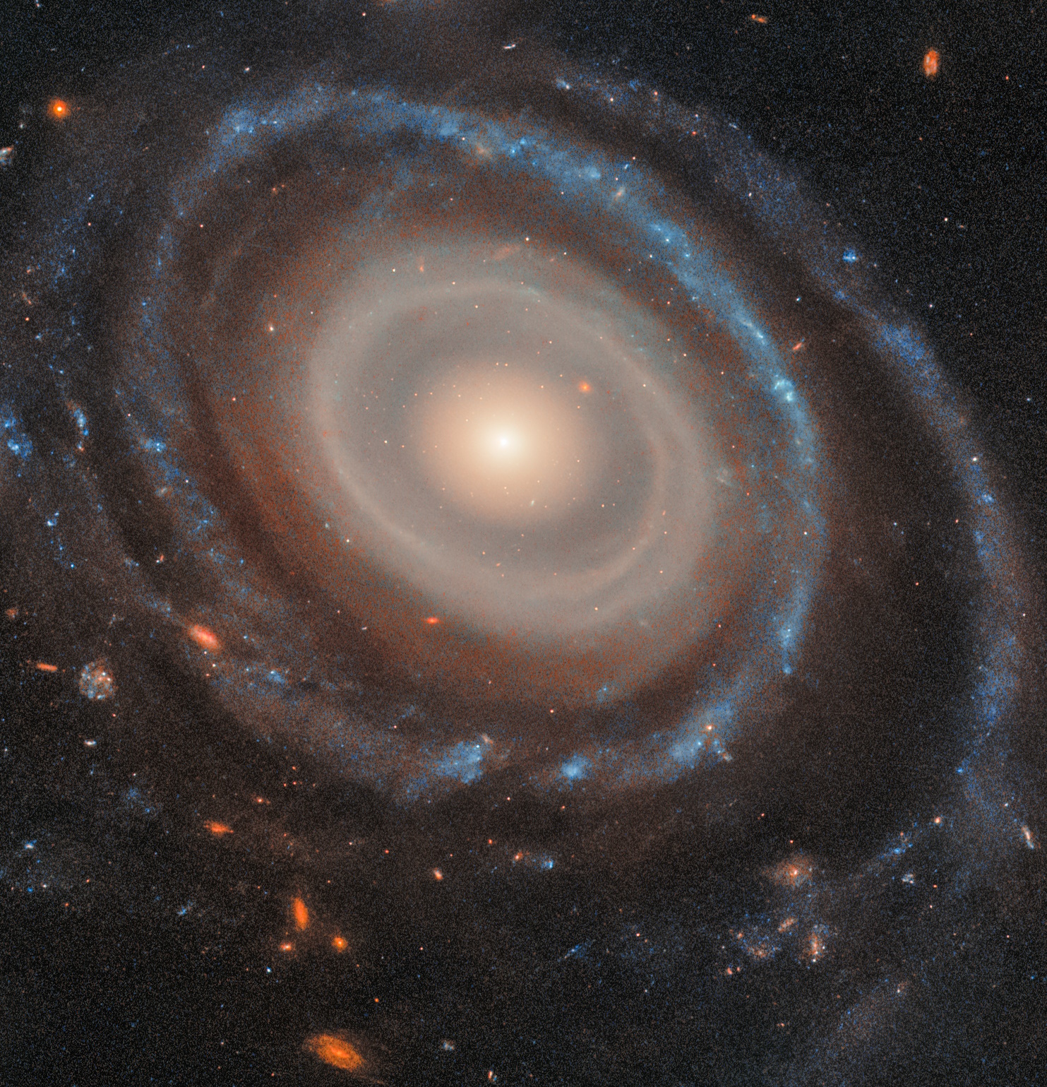 Image of a spiral galaxy with bright core, resembling a bullseye ring galaxy, surrounded by swirling arms containing stars and gas. Background showcases smaller galaxies and scattered stars.
