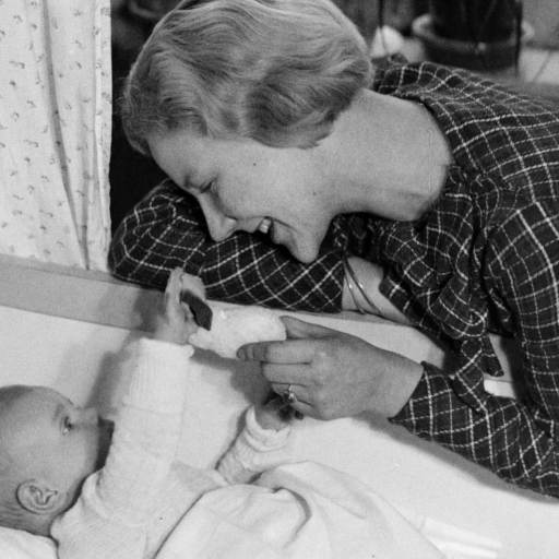 A woman smiles at a baby in a crib, holding a toy while the baby reaches out, attempting their first words. They appear to be in a domestic setting.