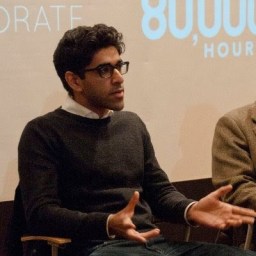 Person with glasses sitting and gesturing with hands, wearing a dark sweater and white shirt. Background features partial text on a wall.