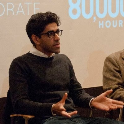 Person with glasses sitting and gesturing with hands, wearing a dark sweater and white shirt. Background features partial text on a wall.