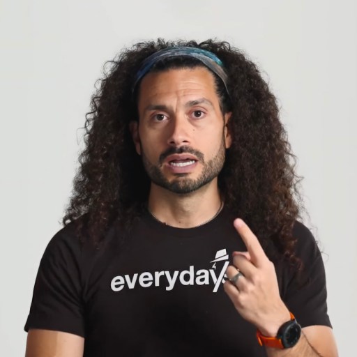 A person with long curly hair and a headband gestures with one hand, wearing a black shirt that says "everyday" and a smartwatch on the other wrist, against a plain background.
