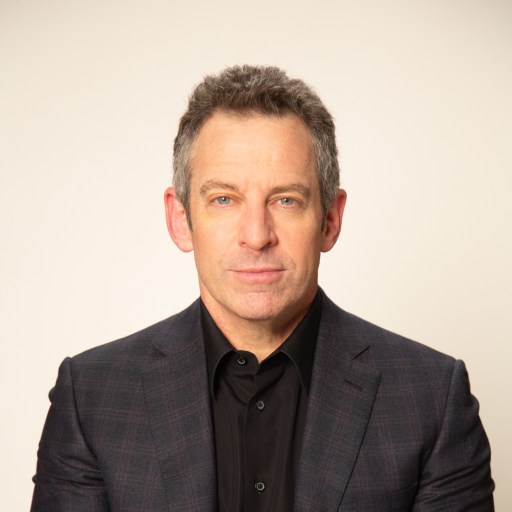 A person in a black shirt and dark blazer poses against a plain background.