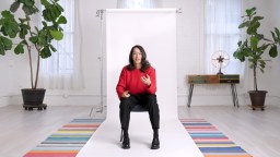 Person in a red sweater sitting on a stool in a bright studio with white walls and colorful striped rugs.