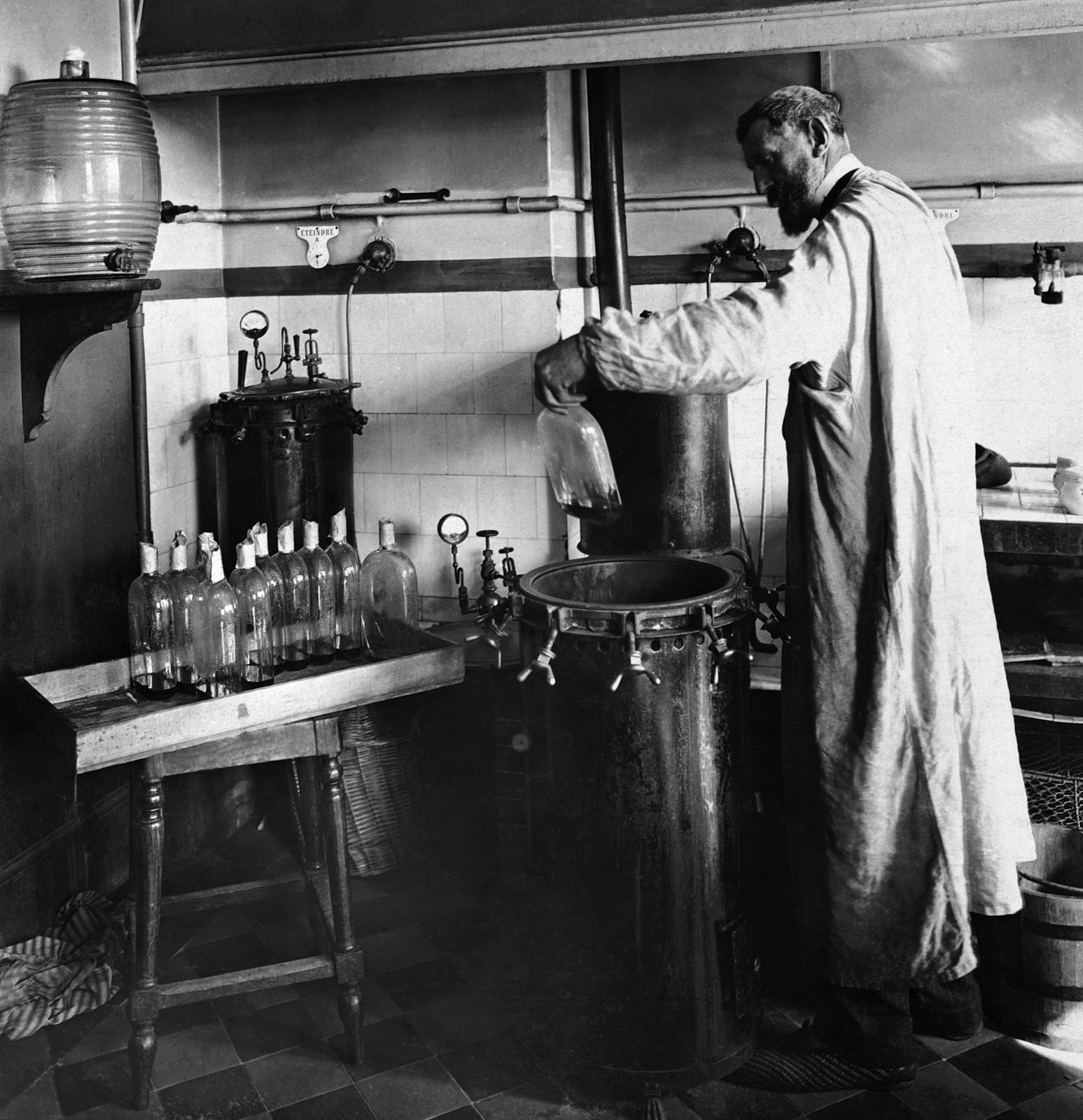 A man in a long coat stands in a laboratory, pouring liquid into a large container, surrounded by glass bottles and laboratory equipment.