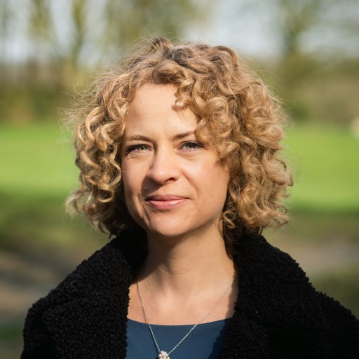 Woman with curly hair wearing a dark jacket and necklace, standing outdoors with trees and grass in the background.