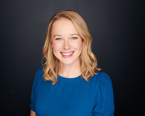 Smiling person with shoulder-length blonde hair, wearing a blue top against a dark background.