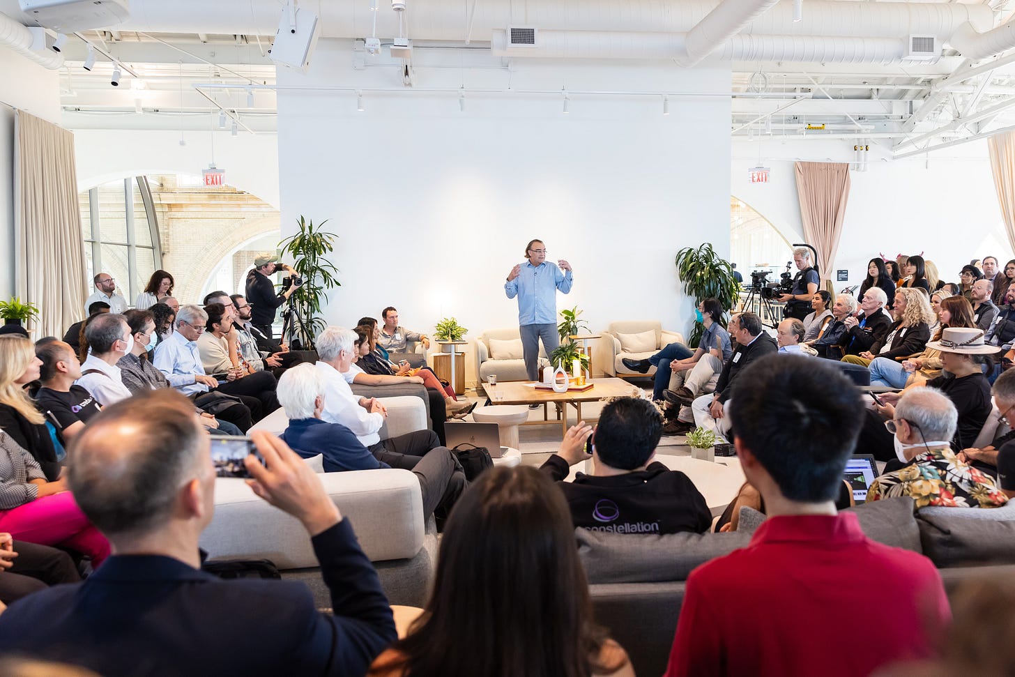 A person speaks to a seated audience in a bright room with plants. Audience members are sitting on couches, some taking photos.