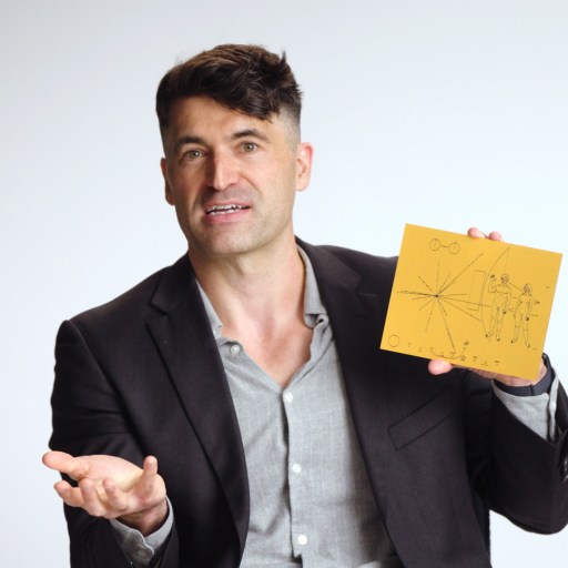 A man in a blazer holds up a yellow card with black line drawings and gestures with his other hand against a plain background.