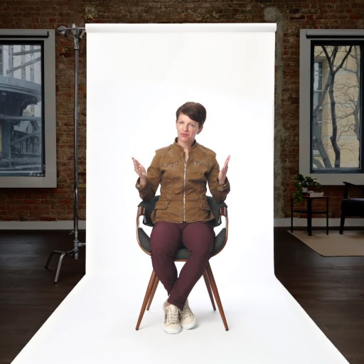 A person sits on a chair in front of a white backdrop in a loft-style room with brick walls, dark flooring, and two windows.