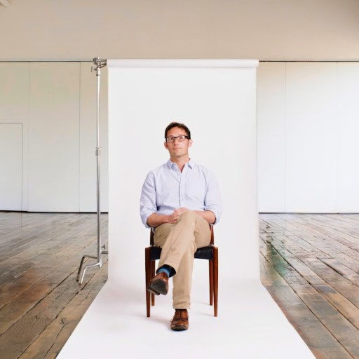 A person sitting on a chair with legs crossed in front of a white backdrop in a spacious room with wooden floors and large windows.