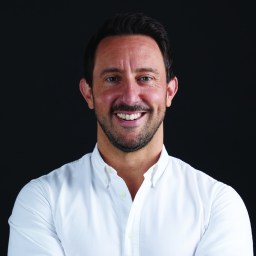 A man with short dark hair and facial hair smiles, wearing a white button-up shirt, against a black background.