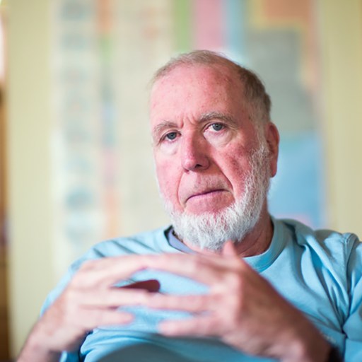 An older man with a white beard sits in a room, wearing a light blue shirt, hands clasped together. Bookshelves and artwork are visible in the background.