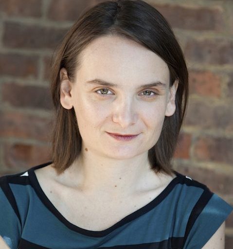 A woman with straight brown hair and light skin wearing a blue and black striped top stands in front of a brick wall, looking directly at the camera.