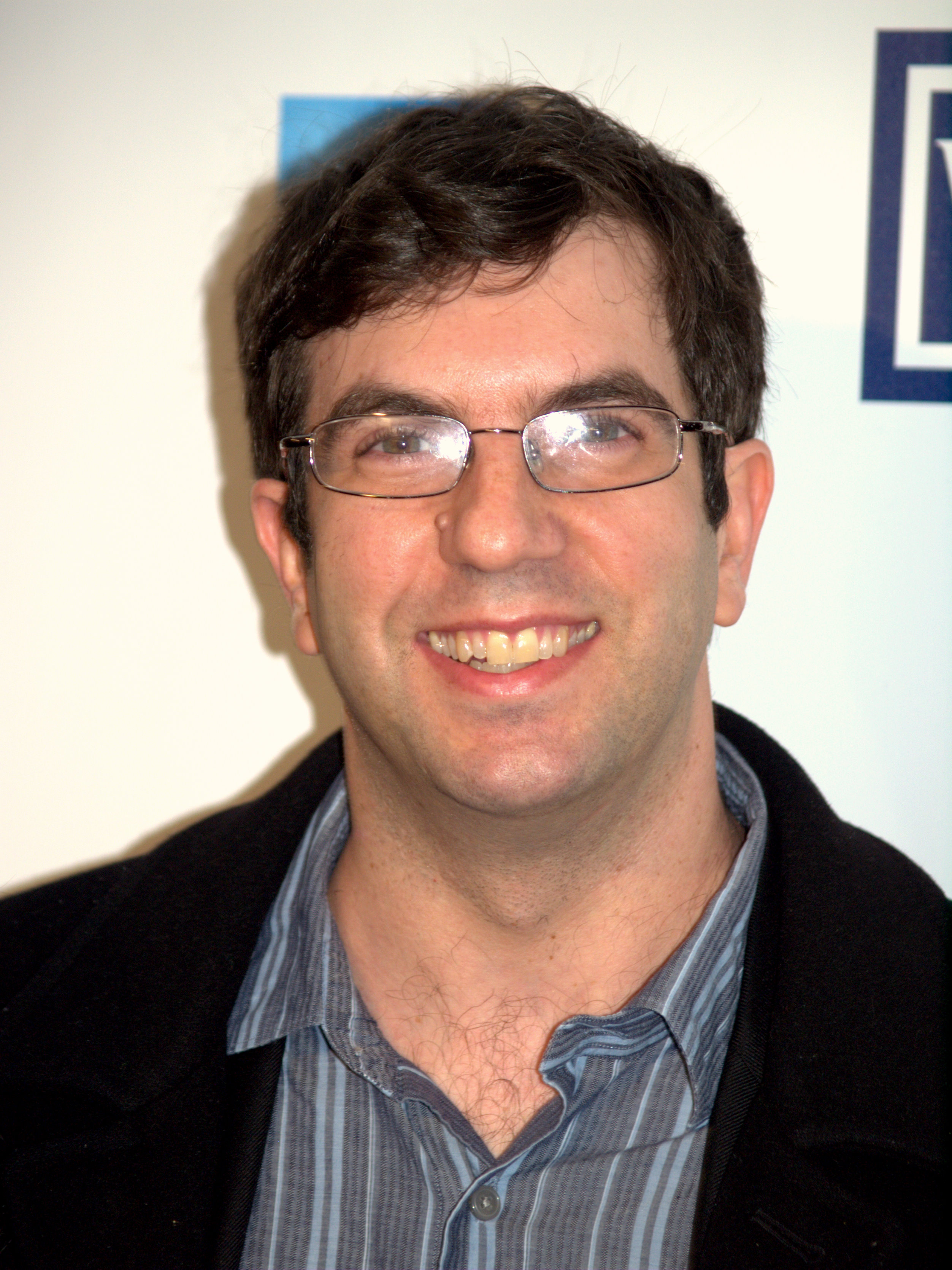 A man with short dark hair and glasses, reminiscent of AJ Jacobs, smiles while wearing a striped shirt and dark jacket against a light-colored backdrop.