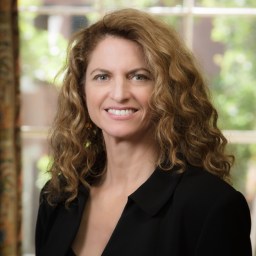 A woman with curly light brown hair, wearing a black blazer, stands indoors in front of a window with greenery visible outside.