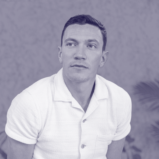 David Perell, in a short-sleeve white shirt, sits in front of a blurred background with plants, looking slightly to the side.