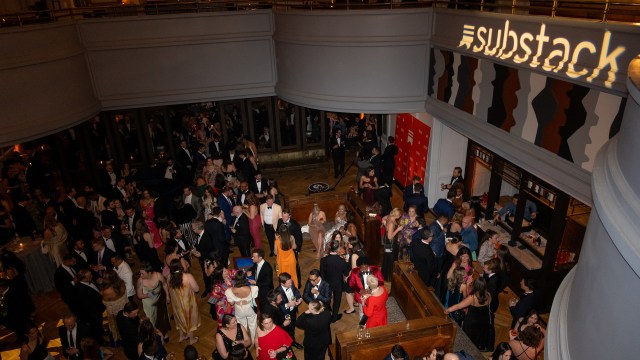 Large crowd of well-dressed people socializing at an indoor event; "Substack" is projected on a wall above.