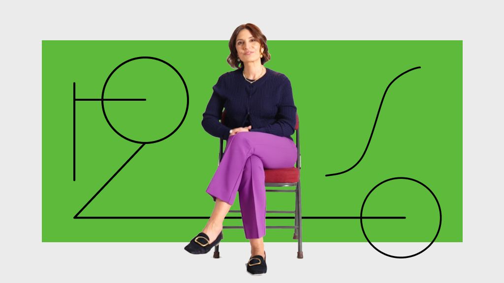 A woman sits on a chair with legs crossed, wearing a navy sweater and purple pants, in front of a green background with black abstract line art.