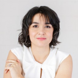 A woman with short dark hair and a white sleeveless top sits with her arms crossed, looking directly at the camera against a plain light background.