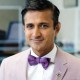 A man wearing a light pink blazer, white shirt, and purple bow tie is smiling slightly while facing the camera.