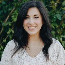 A woman with long dark hair and a white blouse smiles at the camera while standing outdoors in front of green foliage.