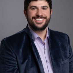 Man with short dark hair and beard, wearing a blue velvet jacket over a light purple shirt, smiles in front of a plain gray background.