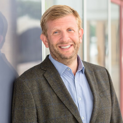 A man with blonde hair and a beard, wearing a gray blazer and light blue shirt, stands smiling in front of a glass wall.