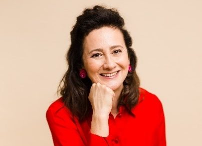 Woman with dark hair wearing a red blouse and black pants sits on a stool, smiling at the camera with her chin resting on her hand against a beige background.