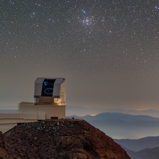 The Vera Rubin Observatory is situated on a rocky hilltop under a clear, star-filled night sky, with distant mountains and a bright planet visible on the horizon, inspiring astronomers to solve puzzles of the universe.