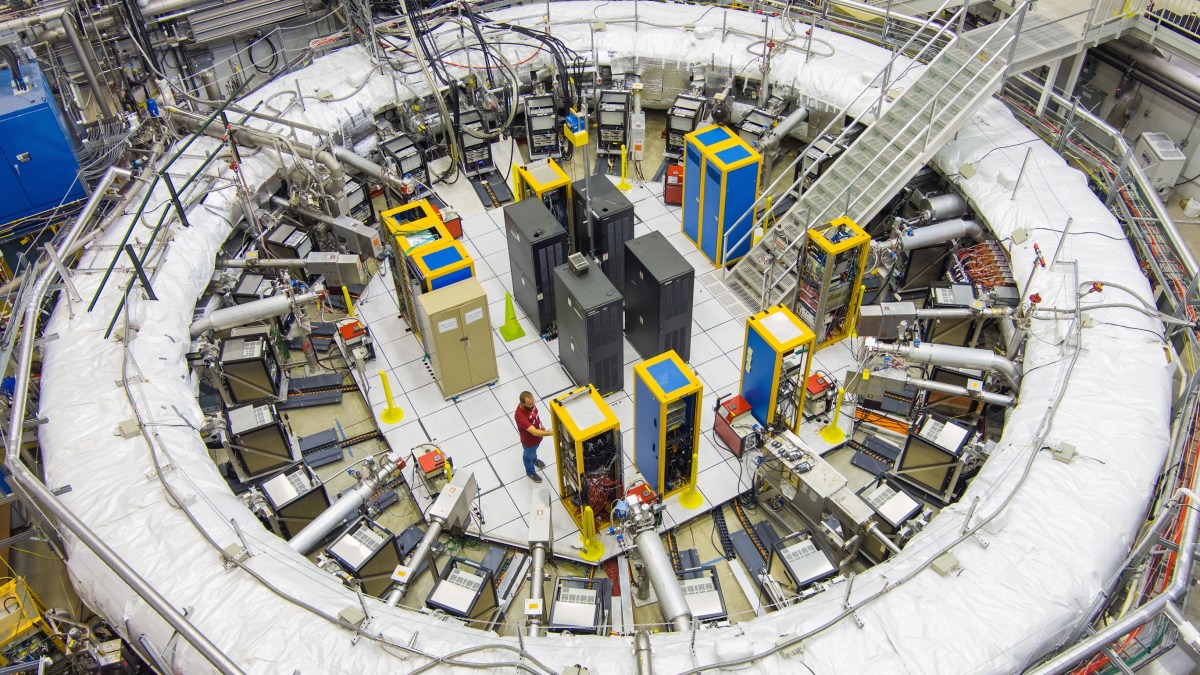 A large circular particle accelerator laboratory with various machines, cables, and equipment; two people are working near the center on experiments related to the muon g-2 anomaly.