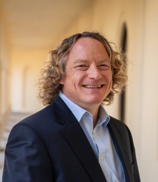 A man with curly blond hair wearing a dark suit and light blue shirt smiles while standing in a sunlit hallway with arched doorways in the background.